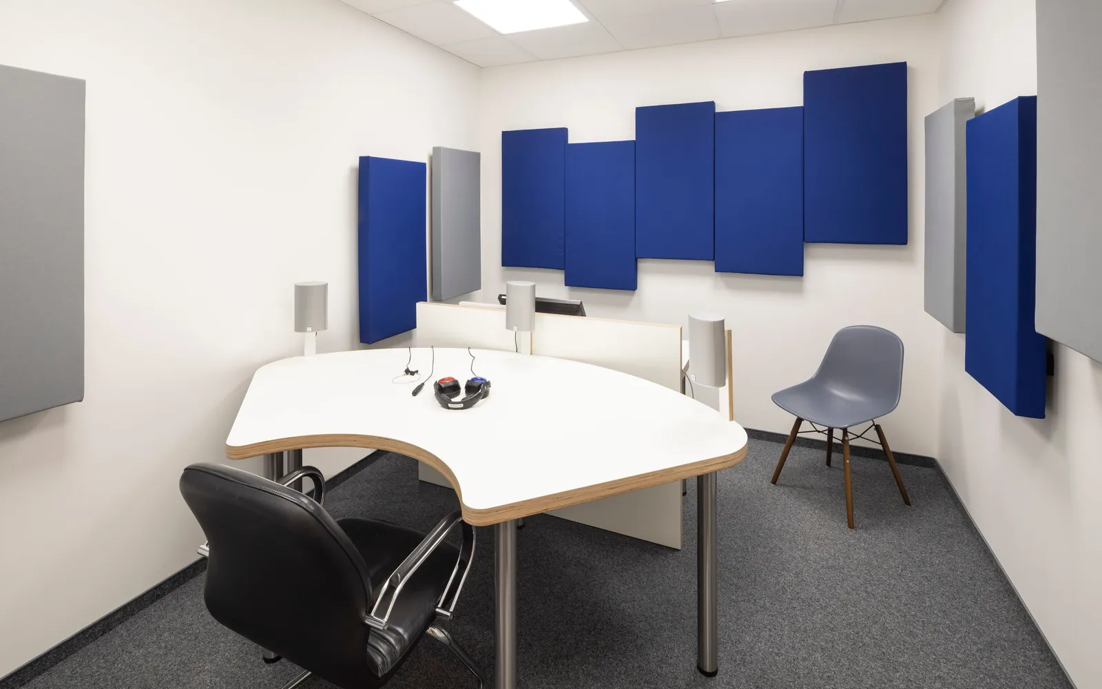 Schallisolierter Audiometrieraum mit blauen und grauen Akustikpaneelen an den Wänden, grauem Teppichboden und einem geschwungenen weißen Tisch mit Kopfhörern und Antworttaster für den Hörtest